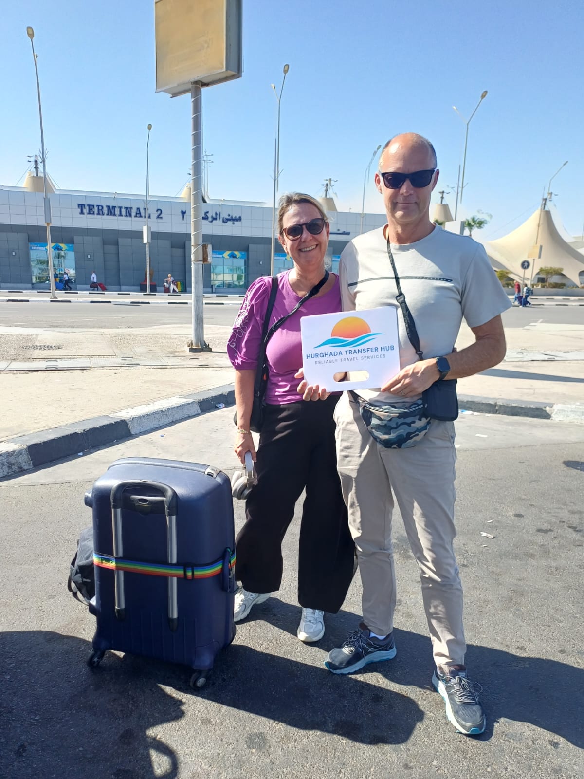 Man and woman posing with a wheeled suitcase and a Hurghada Transfer Hub welcome sign outside Hurghada Airport Terminal 2