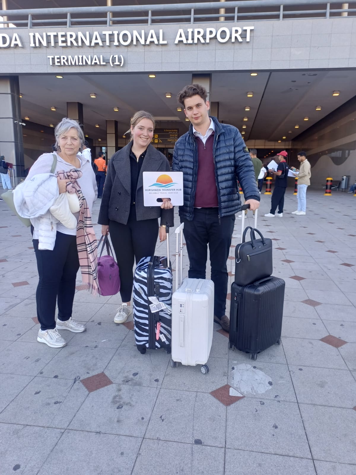 Three travelers with luggage posing with a Hurghada Transfer Hub welcome sign outside Hurghada International Airport Terminal 1