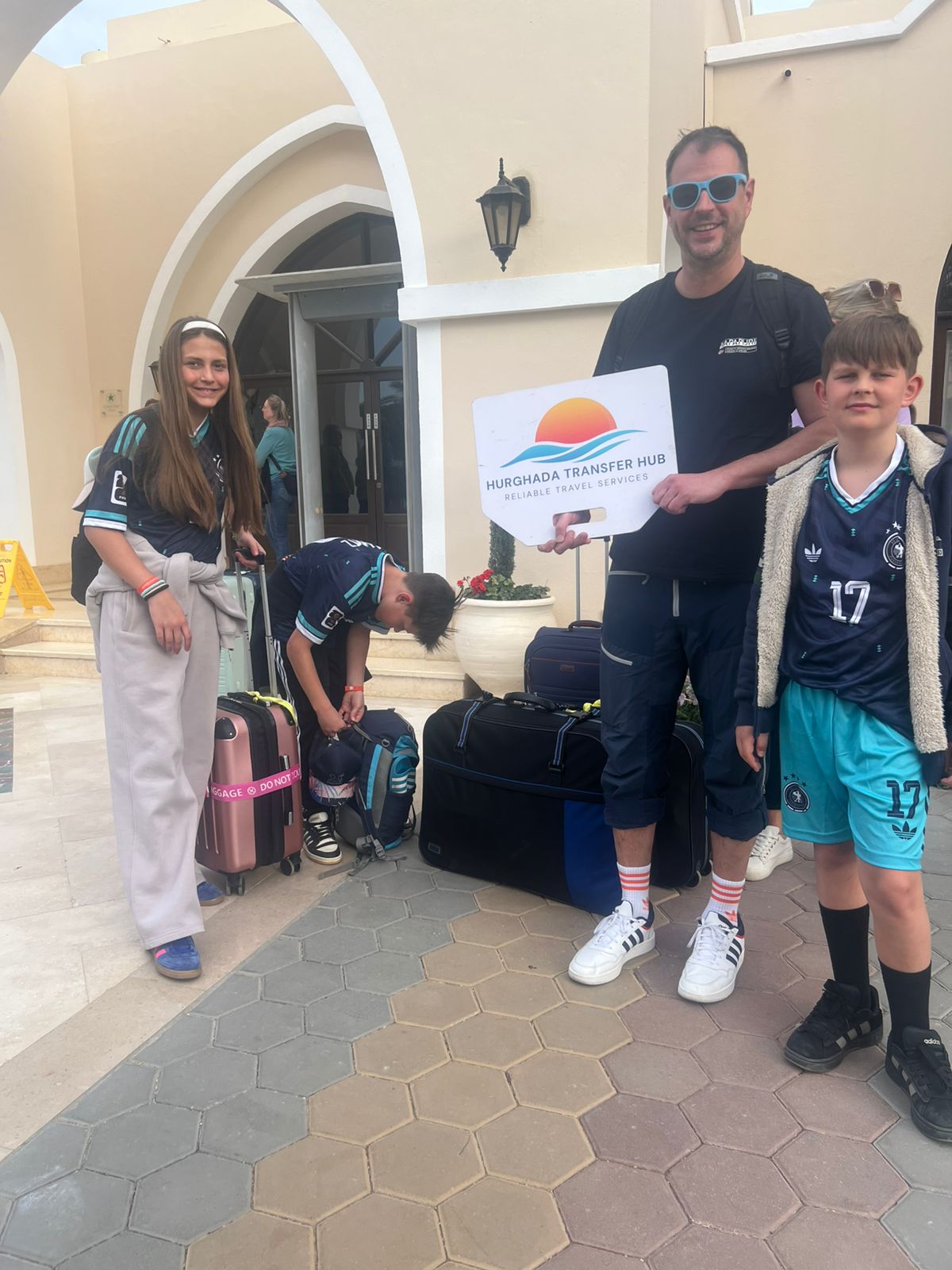 Family of four with luggage posing with a Hurghada Transfer Hub welcome sign outside a resort entrance