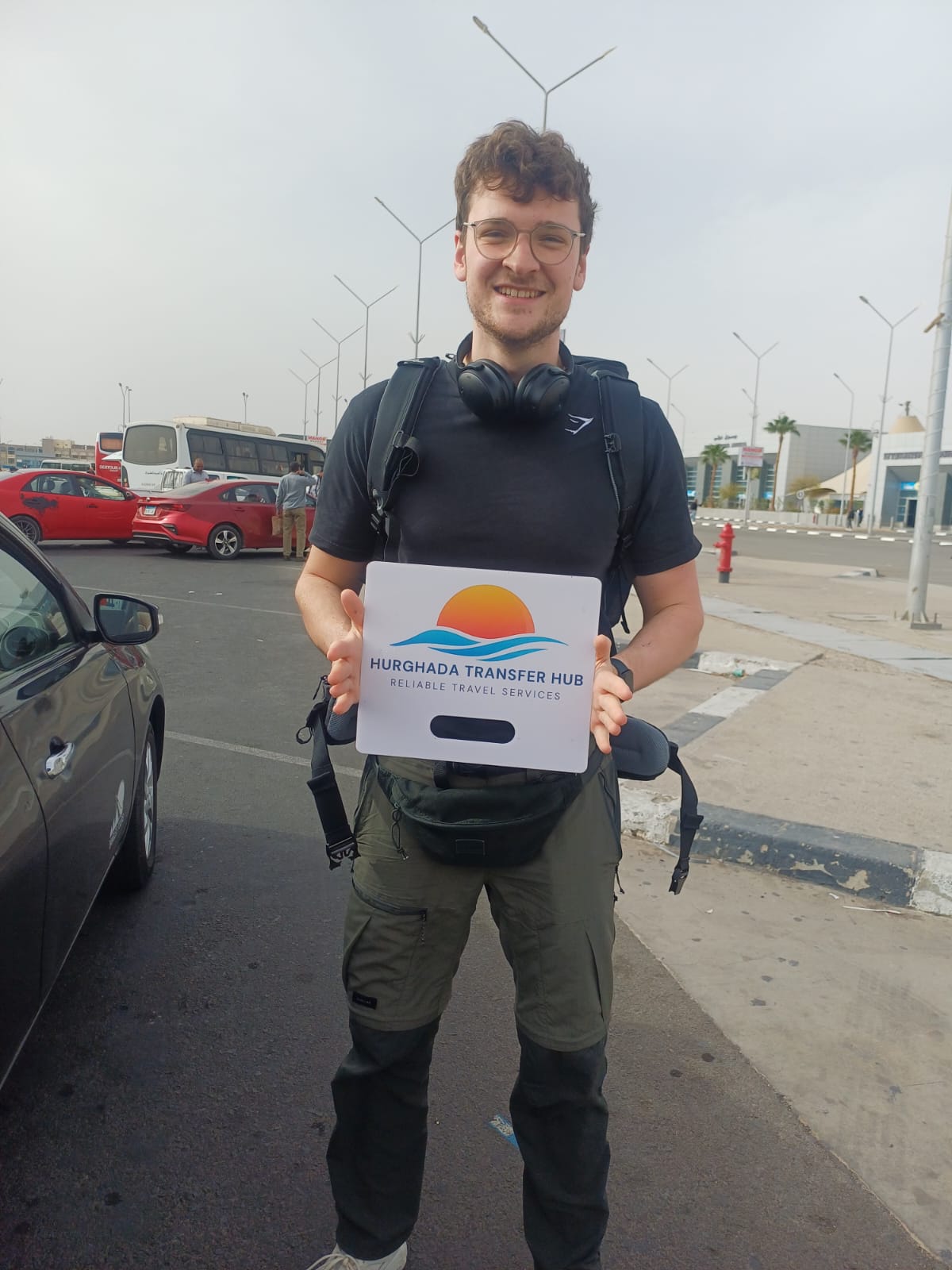 Young man holding a Hurghada Transfer Hub welcome sign in an airport pick-up area with vehicles in the background