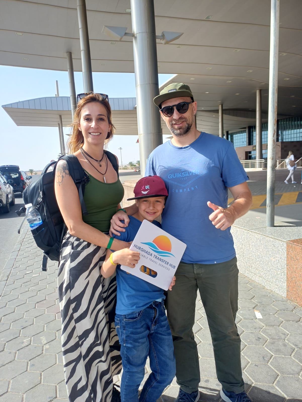 Family of three smiling outside an airport terminal entrance, with the son holding a Hurghada Transfer Hub welcome sign and the father giving a thumbs-up
