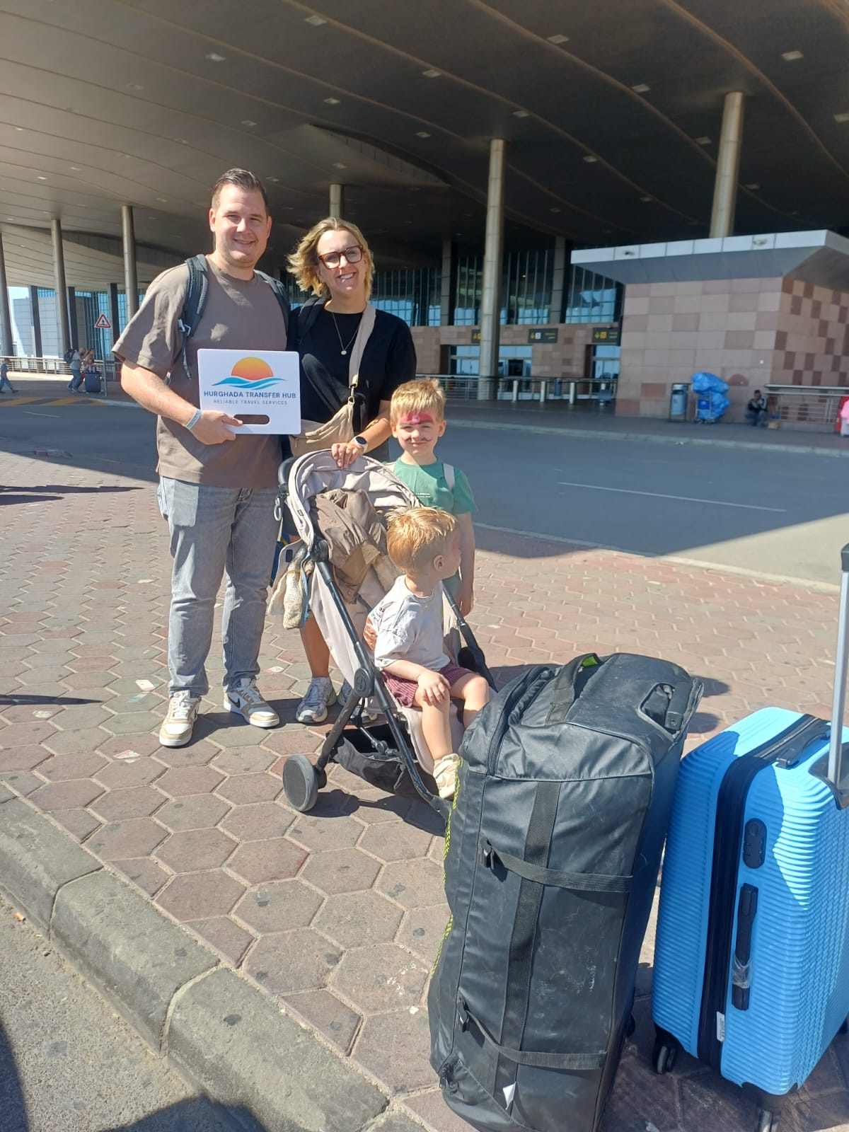 Family of four with luggage posing with a Hurghada Transfer Hub welcome sign outside an airport terminal