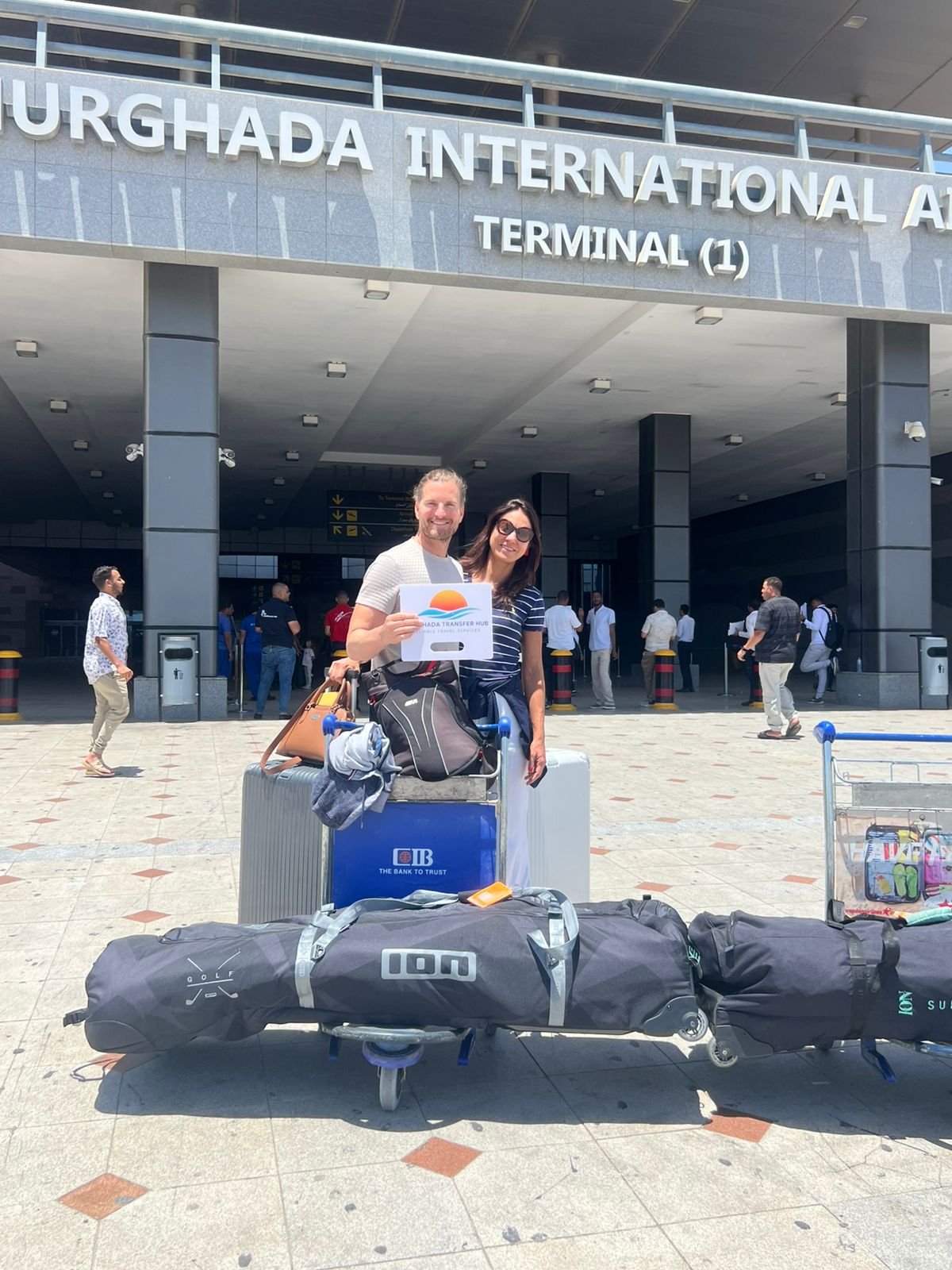 Man and woman with a luggage cart loaded with suitcases, holding a Hurghada Transfer Hub welcome sign outside Hurghada International Airport Terminal 1