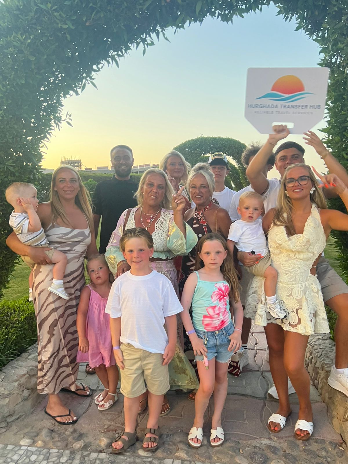 A large multi-generational family smiling outdoors under a resort garden archway, with one member holding a Hurghada Transfer Hub welcome sign