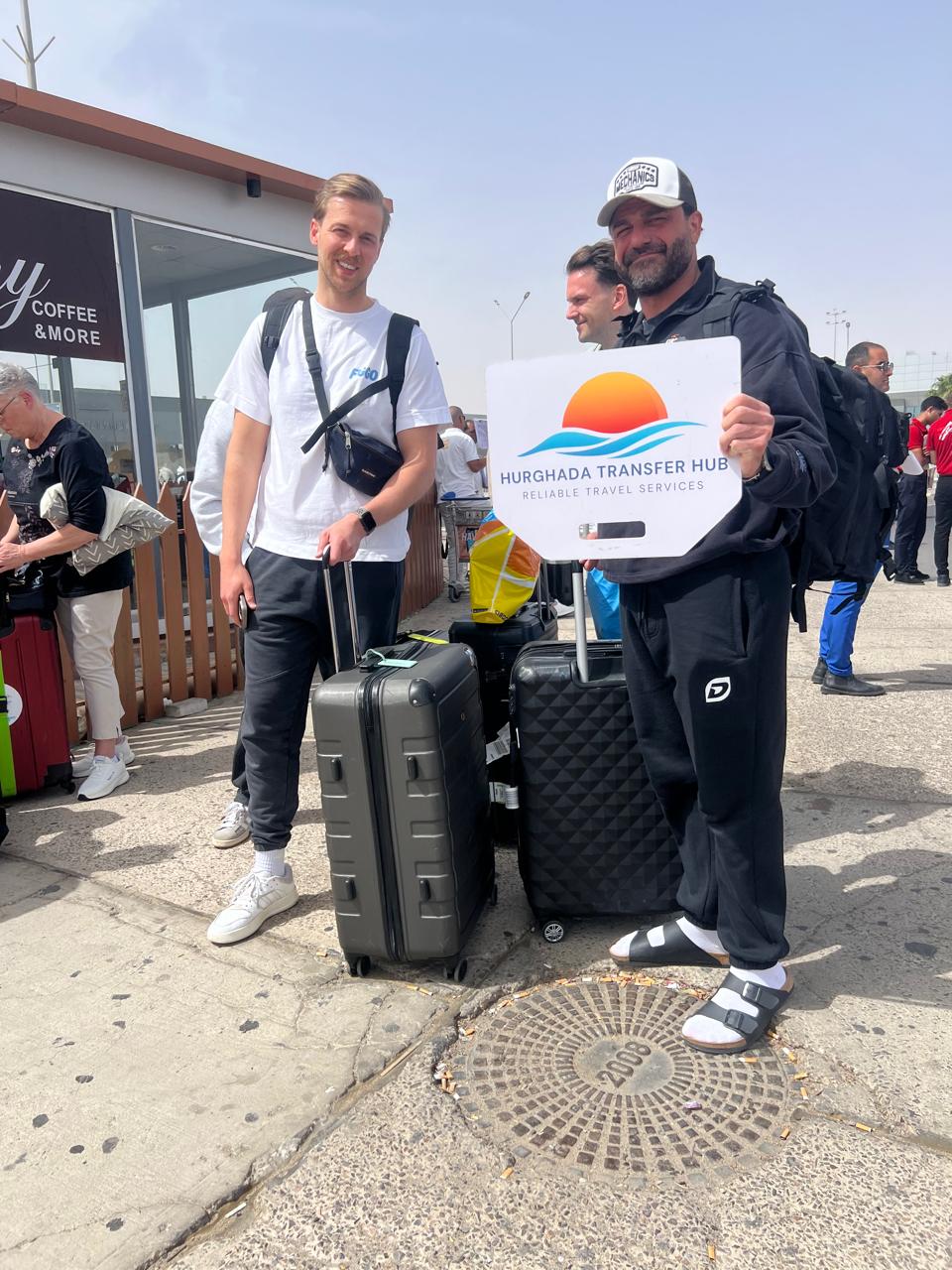 A smiling traveler standing beside a driver holding a Hurghada Transfer Hub welcome sign, with large rolling suitcases outside an airport terminal