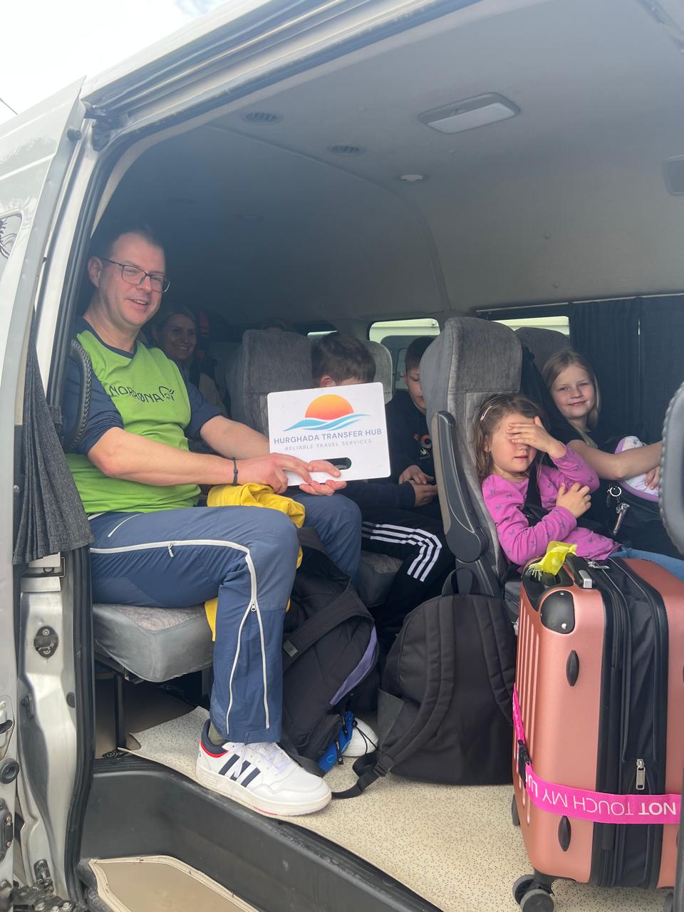 A family seated inside a private passenger transfer van, with the father holding up a white Hurghada Transfer Hub welcome sign next to their luggage