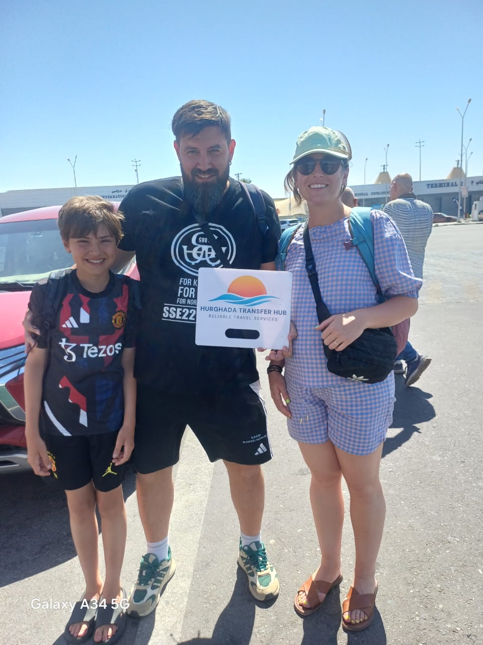 A smiling family of three posing outside Hurghada Airport Terminal 2 next to their transfer vehicle, holding a Hurghada Transfer Hub pickup sign