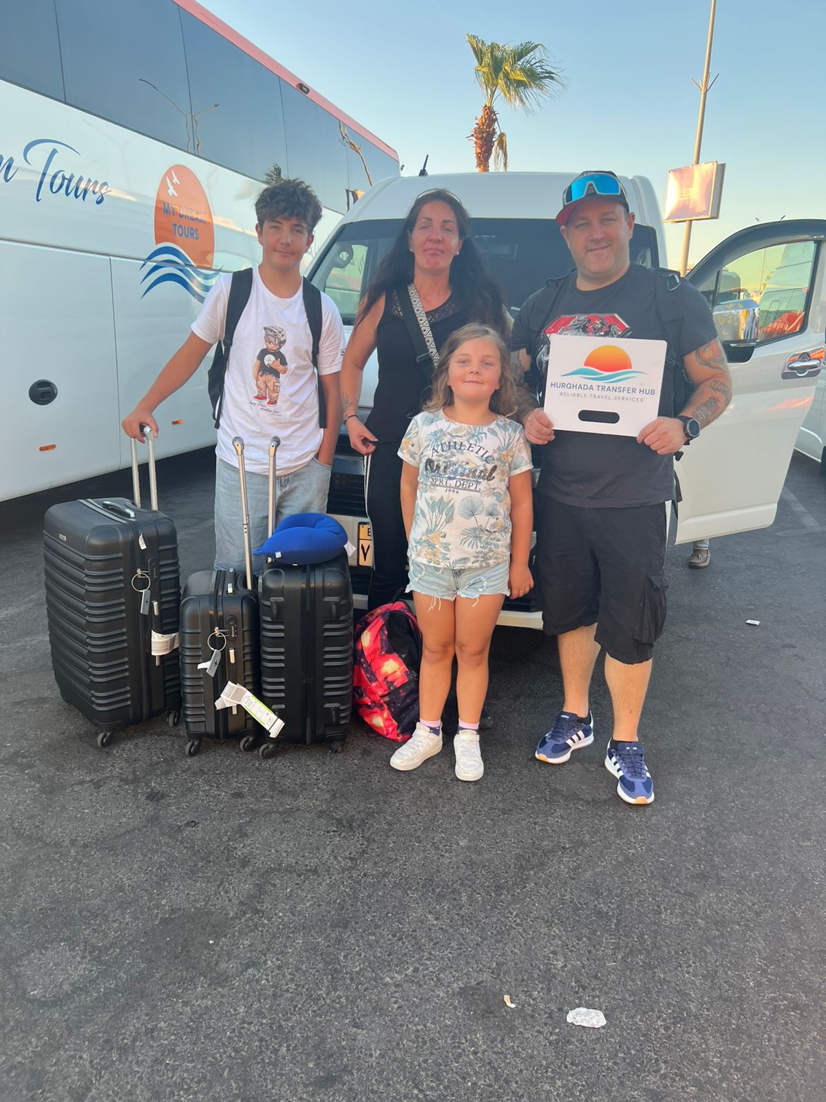 A smiling family of four standing with their luggage in front of a private transfer van, with the father holding a Hurghada Transfer Hub welcome sign