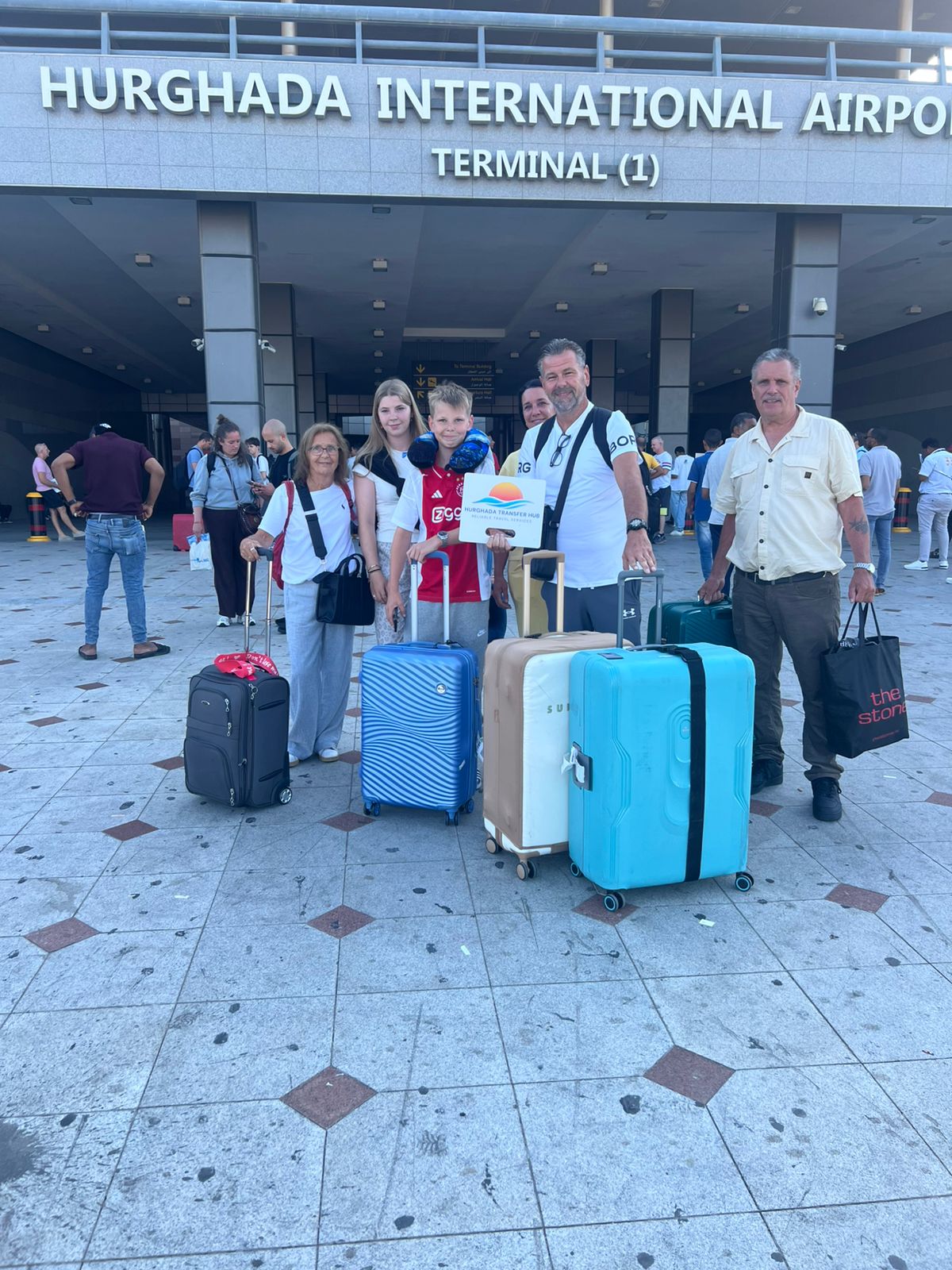 A smiling family of six travelers standing outside Hurghada International Airport Terminal 1 with their rolling luggage, while a young boy holds a Hurghada Transfer Hub welcome sign