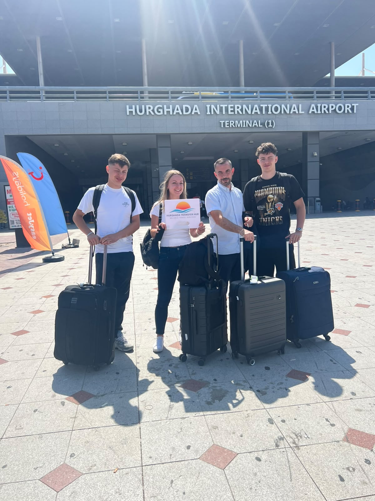 A group of travelers posing with their transfer driver and luggage outside Hurghada International Airport Terminal 1, holding a Hurghada Transfer Hub welcome sign
