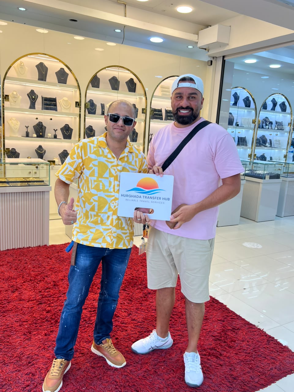 A Hurghada Transfer Hub representative and a smiling traveler posing together holding a company welcome sign inside a resort shopping arcade