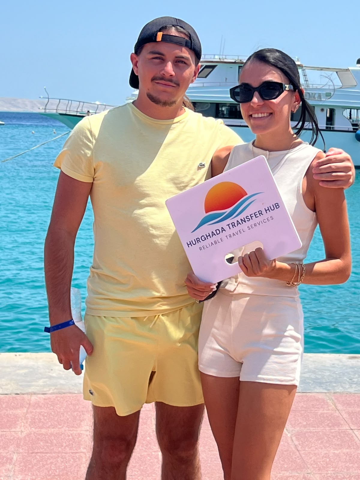 A smiling couple posing at a sunny marina holding a Hurghada Transfer Hub welcome sign with turquoise sea and a boat in the background