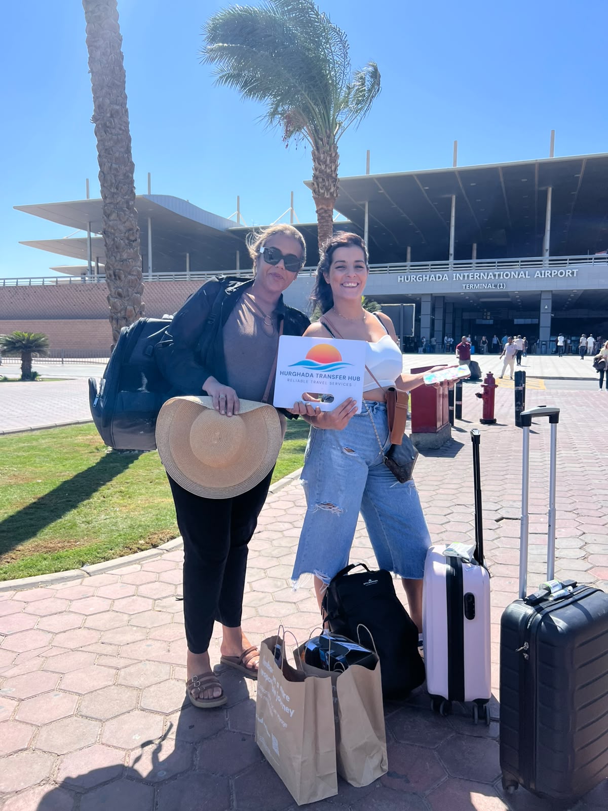 Two smiling female travelers posing outside Hurghada International Airport Terminal 1 holding a Hurghada Transfer Hub welcome sign next to their luggage